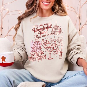Puede incluir: Sudadera color crema con un dise&ntilde;o festivo en rojo. El gr&aacute;fico incluye un &aacute;rbol de Navidad, Pap&aacute; Noel en una copa de Martini y el texto "It's the Most Wonderful Time of the Year". Se sostiene una taza con un hombre de jengibre.