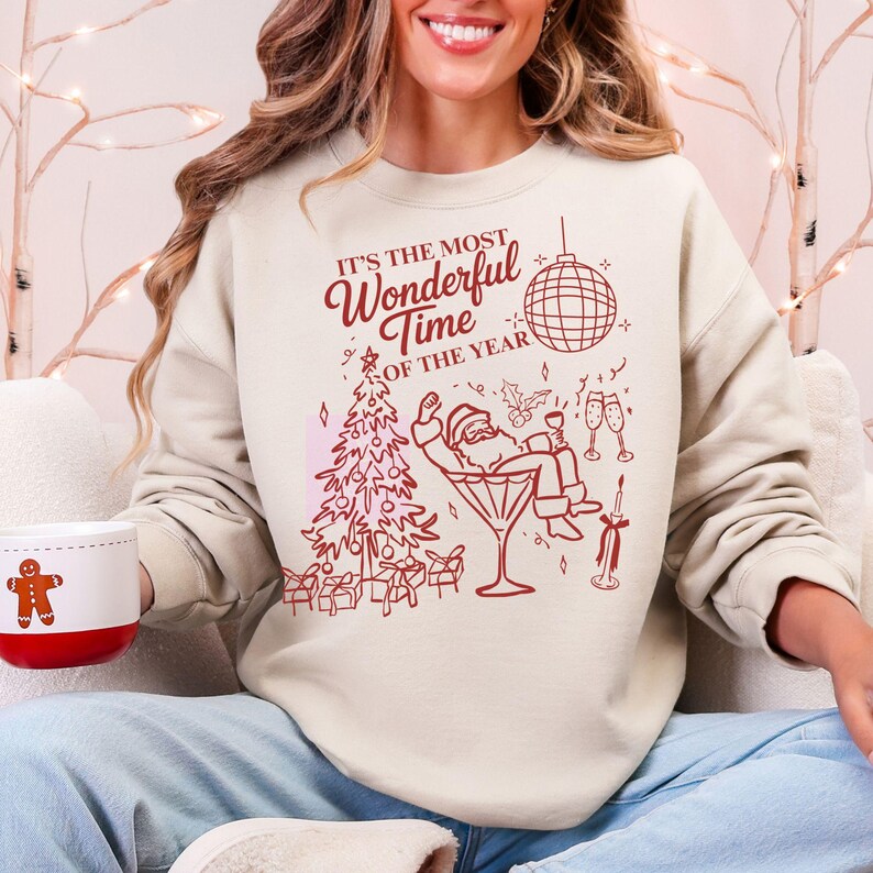 Puede incluir: Sudadera color crema con un dise&ntilde;o festivo en rojo. El gr&aacute;fico incluye un &aacute;rbol de Navidad, Pap&aacute; Noel en una copa de Martini y el texto "It's the Most Wonderful Time of the Year". Se sostiene una taza con un hombre de jengibre.