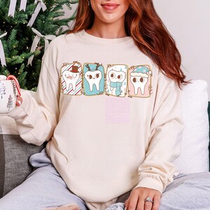 May include: Cream-colored long-sleeve shirt featuring four cartoon teeth in festive holiday attire. Each tooth is inside a rectangular frame with decorative elements like a Santa hat, reindeer antlers, a scarf, and a winter hat. The shirt is worn by a person.
