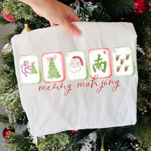 May include: A white t-shirt featuring a graphic of five mahjong tiles with Christmas-themed illustrations including a dragon, a pine tree, Santa Claus, a green Chinese character, and pinecones, with the red text merry mahjong printed below.