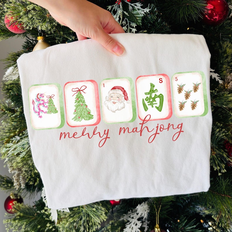 May include: A white t-shirt featuring a graphic of five mahjong tiles with Christmas-themed illustrations including a dragon, a pine tree, Santa Claus, a green Chinese character, and pinecones, with the red text merry mahjong printed below.