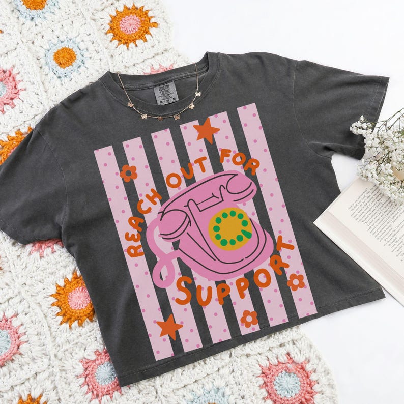 May include: Dark gray t-shirt with a pink and white graphic featuring a pink rotary phone and the words "REACH OUT FOR SUPPORT". The shirt has a crew neck and short sleeves. A gold necklace with butterfly charms is visible.