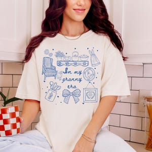 May include: Cream-colored t-shirt with a blue graphic design featuring the text "In my granny era" and various illustrations. The illustrations include a chair, cat, dress, and kitchen items.