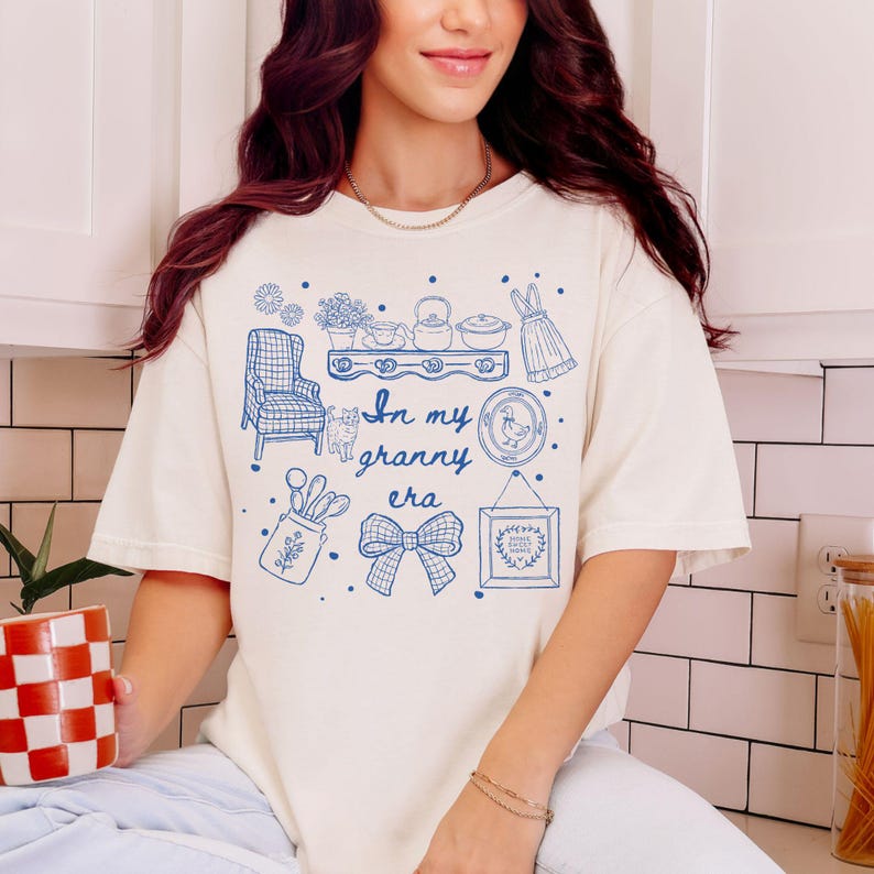 May include: Cream-colored t-shirt with a blue graphic design featuring the text "In my granny era" and various illustrations. The illustrations include a chair, cat, dress, and kitchen items.