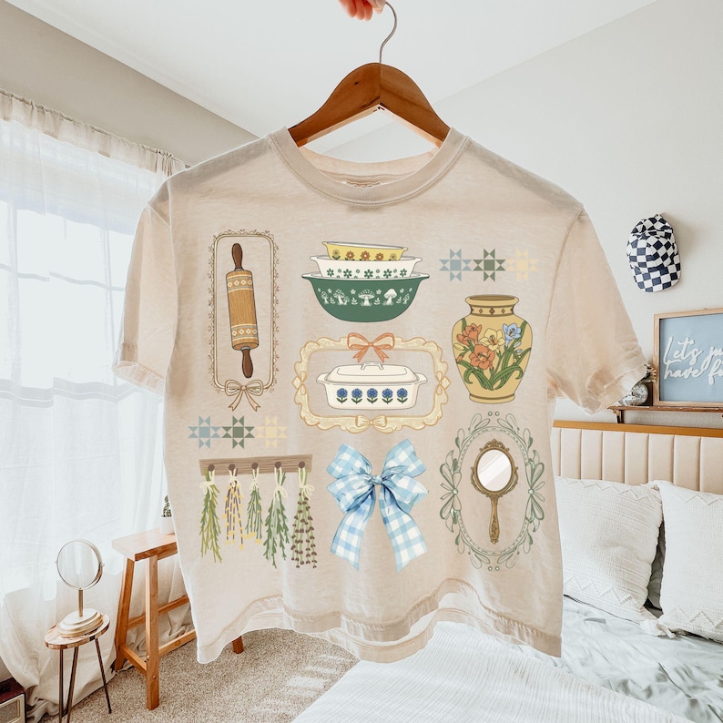 May include: Cream-colored t-shirt featuring a collection of vintage kitchen-themed illustrations. The design includes a rolling pin, bowls, a vase with flowers, a mirror, and a blue gingham bow. The shirt is hanging on a wooden hanger.