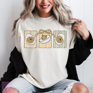 May include: Cream-colored sweatshirt featuring a graphic of three deviled eggs. The eggs are in decorative frames with vertical stripes and a bow. The design includes brown, white, and tan colors. The sweatshirt is a casual, everyday garment.