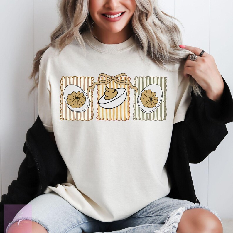 May include: Cream-colored sweatshirt featuring a graphic of three deviled eggs. The eggs are in decorative frames with vertical stripes and a bow. The design includes brown, white, and tan colors. The sweatshirt is a casual, everyday garment.