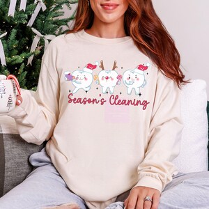 Puede incluir: Una camiseta de manga larga color crema con un gr&aacute;fico de tres dientes de dibujos animados con gorros de Pap&aacute; Noel y objetos navide&ntilde;os. El texto "Season's Cleaning" est&aacute; escrito en cursiva debajo. La camiseta es de estilo c&oacute;modo e informal.