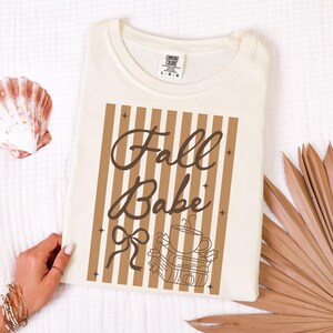 May include: Cream-colored sweatshirt with the words "Fall Babe" in brown script, set against a background of vertical brown and cream stripes. A graphic of a coffee cup, books, and a pumpkin is below the text. The brand tag is visible.