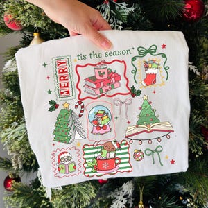 Może przedstawiać: Biała bluza z motywem świątecznym z kolorowymi ilustracjami. Zawiera choinkę, książki, kulę śnieżną i kubek z napisem "tis the season" i "MERRY". Projekt zawiera czerwone, zielone i r&oacute;żowe akcenty.