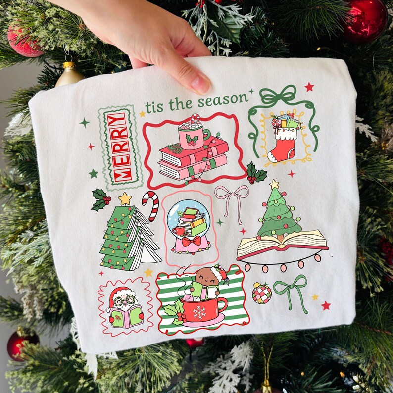 Może przedstawiać: Biała bluza z motywem świątecznym z kolorowymi ilustracjami. Zawiera choinkę, książki, kulę śnieżną i kubek z napisem "tis the season" i "MERRY". Projekt zawiera czerwone, zielone i r&oacute;żowe akcenty.
