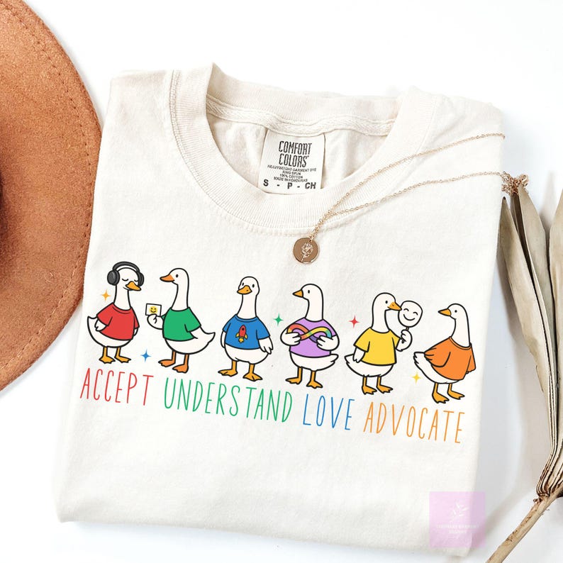 May include: Cream-colored t-shirt featuring a line of cartoon ducks, each wearing a different colored shirt and accessories. The text "ACCEPT UNDERSTAND LOVE ADVOCATE" is printed below the ducks in rainbow colors. A gold necklace is draped across the shirt.