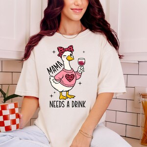 Peut inclure: T-shirt cr&egrave;me avec un dessin d'oie de dessin anim&eacute; portant un pull rose avec un c&oelig;ur et un n&oelig;ud l&eacute;opard. L'oie tient un verre de vin. Le texte "MAMA NEEDS A DRINK" est imprim&eacute; sur le t-shirt.