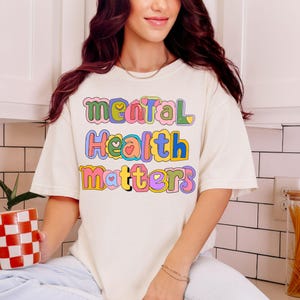 May include: Cream-colored t-shirt with the words "Mental Health Matters" in colorful, bubble letters. The shirt has a relaxed fit and short sleeves. The text is in a playful, retro style, with each word in a different color.