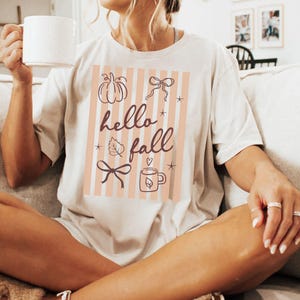 May include: Cream-colored t-shirt with a fall-themed graphic. The design features a pumpkin, bows, a leaf, a mug, and the words "hello fall" in a script font, set against a striped background. The shirt is a casual, comfortable style.