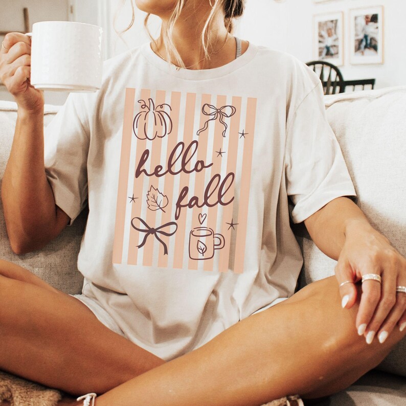May include: Cream-colored t-shirt with a fall-themed graphic. The design features a pumpkin, bows, a leaf, a mug, and the words "hello fall" in a script font, set against a striped background. The shirt is a casual, comfortable style.