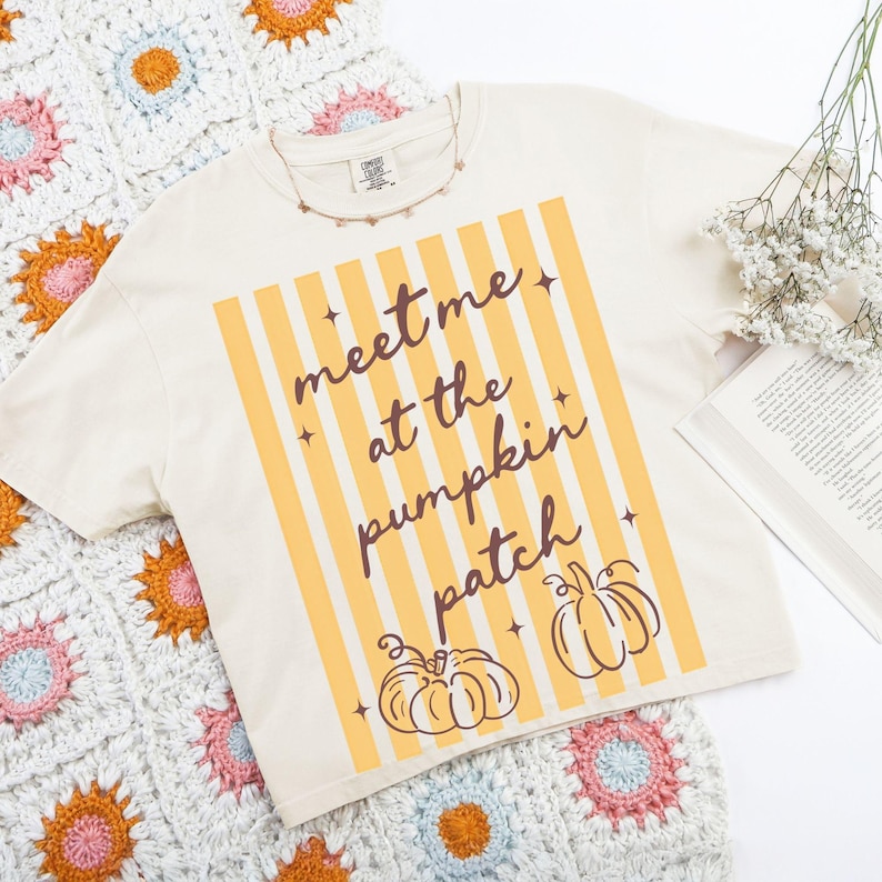 May include: Cream-colored t-shirt with the text "meet me at the pumpkin patch" in a vertical striped design. Two pumpkin illustrations are at the bottom. A crochet blanket with floral patterns and a book are in the background.