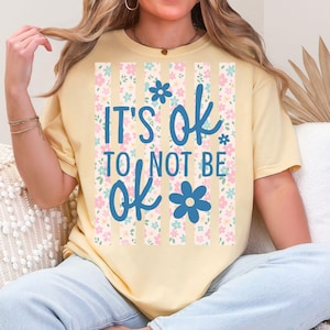 May include: A light yellow t-shirt with the text "IT'S OK TO NOT BE ok" in blue script. The text is set against a vertical striped background with pink and blue floral patterns. The shirt has short sleeves.