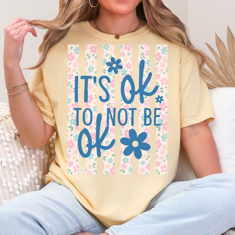 May include: A light yellow t-shirt with the text "IT'S OK TO NOT BE ok" in blue script. The text is set against a vertical striped background with pink and blue floral patterns. The shirt has short sleeves.