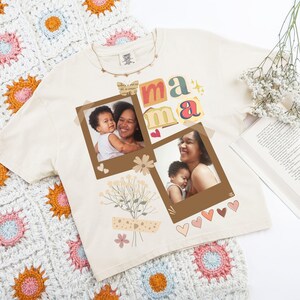 May include: Cream-coloured t-shirt with the word "mama" in colourful letters and two photo prints. The shirt is decorated with floral and heart graphics. A crocheted blanket is in the background.