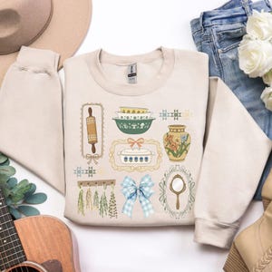 May include: Beige crewneck sweatshirt featuring a collection of vintage kitchen-themed illustrations. The design includes a rolling pin, bowls, a casserole dish, a vase with flowers, a mirror, and a blue gingham bow. The illustrations are framed and detailed.