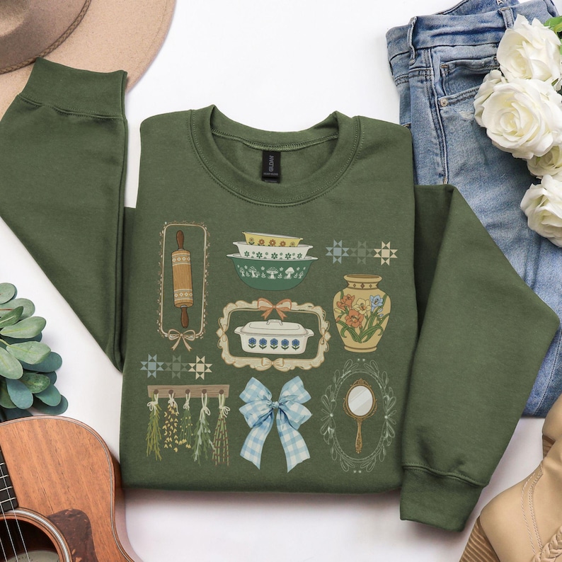 May include: Olive green sweatshirt featuring a collection of vintage kitchen-themed illustrations. The design includes a rolling pin, bowls, a casserole dish, a vase with flowers, a mirror, and a blue gingham bow. The sweatshirt is a comfortable, casual style.