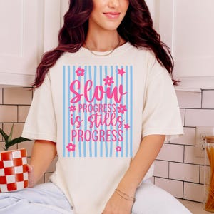 May include: Cream-colored t-shirt with a graphic that reads "Slow Progress is Still Progress" in pink lettering. The graphic is set against a blue and white striped background, adorned with pink flowers and stars.