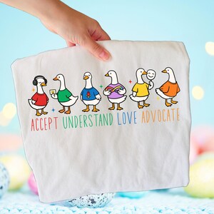 May include: White t-shirt featuring six cartoon ducks, each wearing a different colored shirt and accessories. The text "ACCEPT UNDERSTAND LOVE ADVOCATE" is printed below the ducks in rainbow colors. The shirt is held up by a hand.