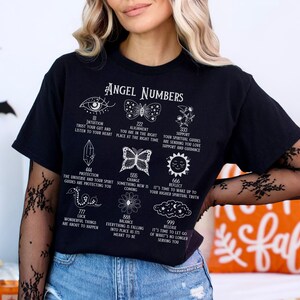 以下が含まれることがあります： 「Angel Numbers」の文字と様々なシンボルと数字がプリントされた黒いTシャツ。目、蝶、花、その他のスピリチュアルなシンボルのイラストが特徴です。テキストには「Intuition」や「Protection」などのフレーズが含まれています。