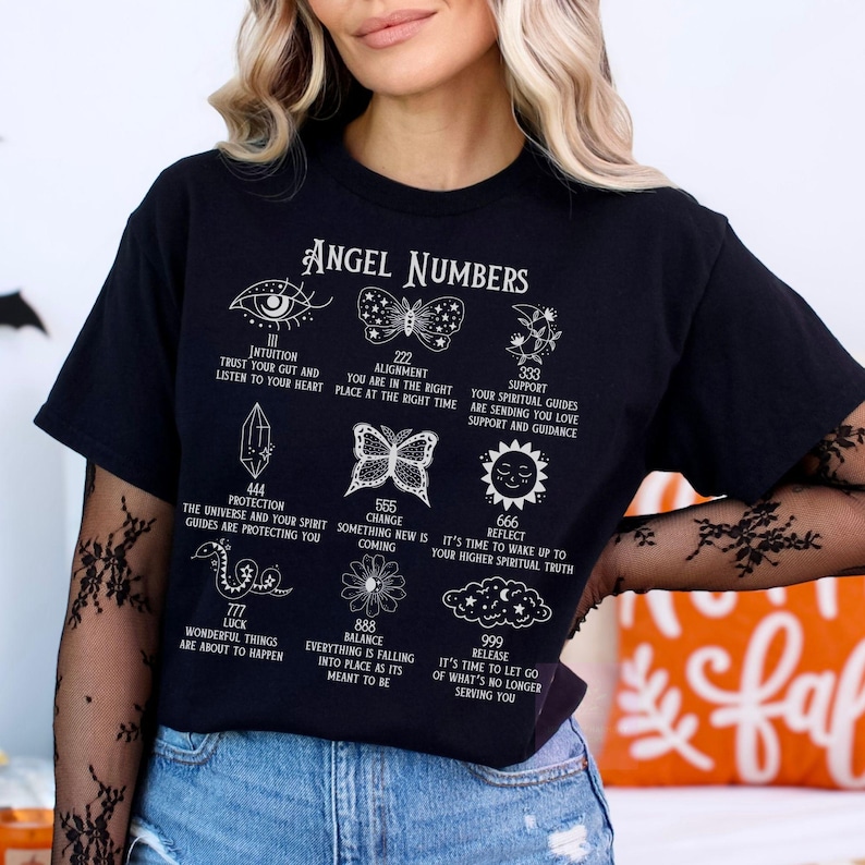 以下が含まれることがあります： 「Angel Numbers」の文字と様々なシンボルと数字がプリントされた黒いTシャツ。目、蝶、花、その他のスピリチュアルなシンボルのイラストが特徴です。テキストには「Intuition」や「Protection」などのフレーズが含まれています。