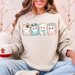 May include: A beige sweatshirt featuring four cartoon teeth, each with a festive design. One tooth wears a hat, another antlers, a third a scarf, and the last a winter hat. The sweatshirt is paired with blue jeans.