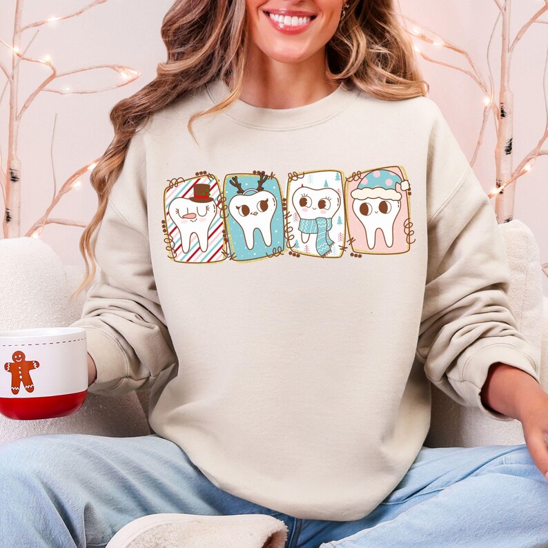 May include: A beige sweatshirt featuring four cartoon teeth, each with a festive design. One tooth wears a hat, another antlers, a third a scarf, and the last a winter hat. The sweatshirt is paired with blue jeans.