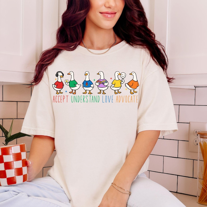 May include: Cream-colored t-shirt featuring a row of cartoon ducks with various accessories and clothing. The ducks are in different colors. Below the ducks is the text "ACCEPT UNDERSTAND LOVE ADVOCATE" in rainbow colors.