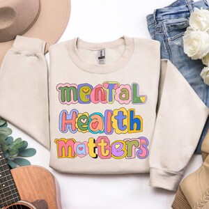 May include: A beige sweatshirt with the words "Mental Health Matters" in colorful, bubble letters. The text includes hearts and is in shades of pink, blue, yellow, and green. The sweatshirt is folded with jeans and a hat.
