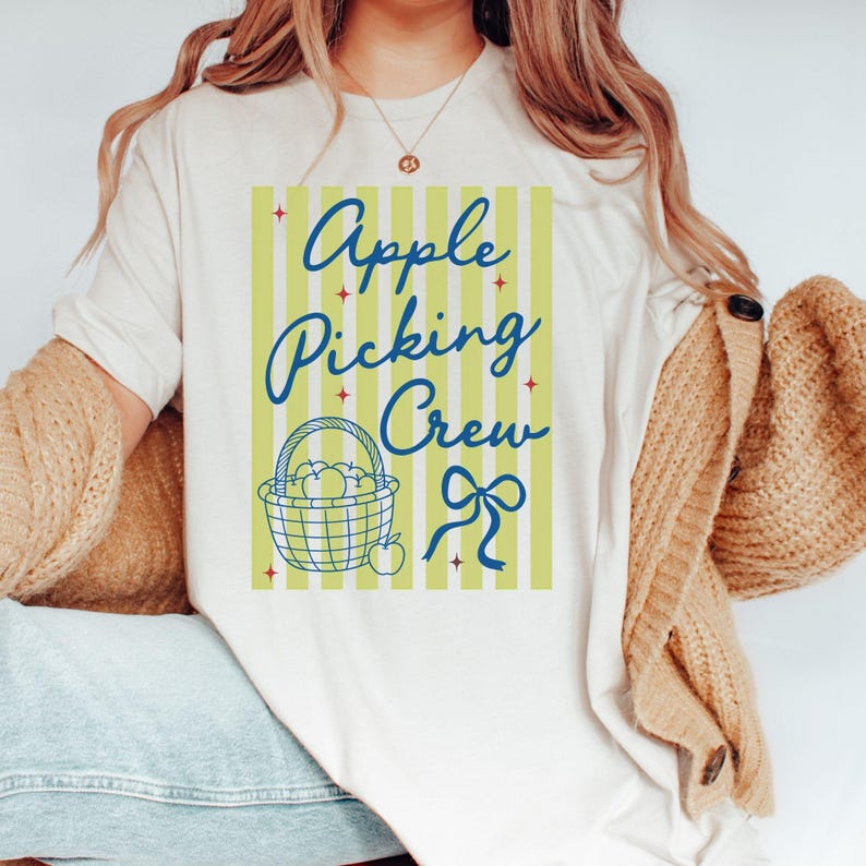May include: Cream-colored t-shirt with the text "Apple Picking Crew" in blue script. The design includes a basket of apples and a bow on a light green striped background. The shirt is part of a fall-themed collection.