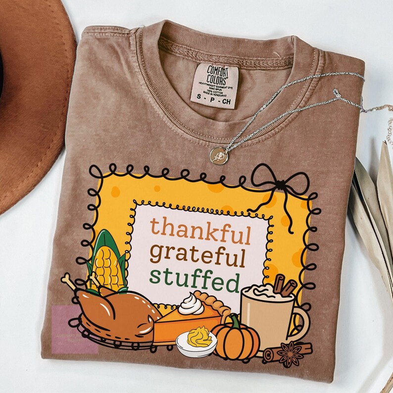 May include: Brown Comfort Colors t-shirt with a graphic that reads "thankful grateful stuffed." The graphic includes a turkey, pumpkin, pie, corn, and a mug of coffee. A silver necklace is draped across the shirt.