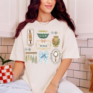 May include: Cream-colored t-shirt featuring a collection of vintage kitchen-themed illustrations. The design includes a rolling pin, bowls, a vase with flowers, a casserole dish, a blue gingham bow, and a hand mirror. The shirt has short sleeves and a relaxed fit.