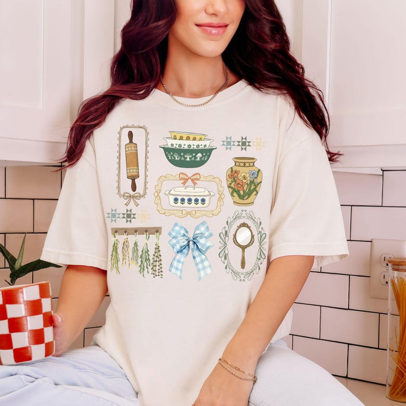 May include: Cream-colored t-shirt featuring a collection of vintage kitchen-themed illustrations. The design includes a rolling pin, bowls, a vase with flowers, a casserole dish, a blue gingham bow, and a hand mirror. The shirt has short sleeves and a relaxed fit.