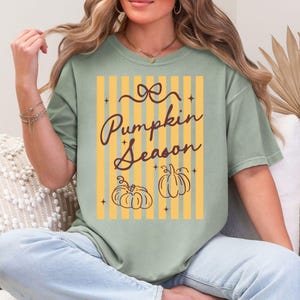 May include: Sage green t-shirt with the text "Pumpkin Season" in a script font. The design includes two pumpkin illustrations and a background of yellow and orange vertical stripes. The shirt is a relaxed fit.