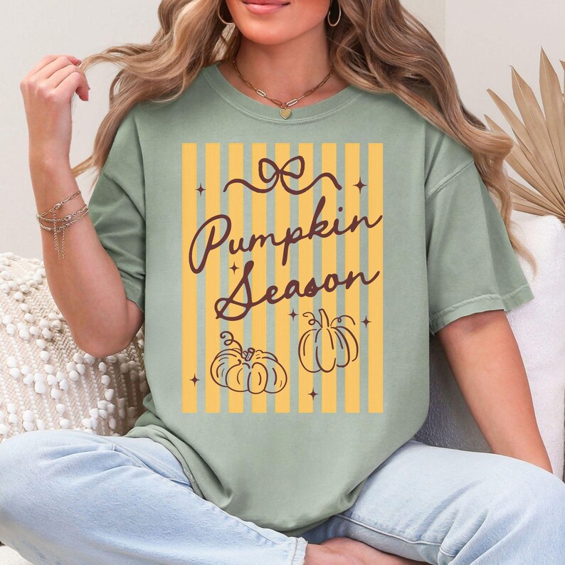 May include: Sage green t-shirt with the text "Pumpkin Season" in a script font. The design includes two pumpkin illustrations and a background of yellow and orange vertical stripes. The shirt is a relaxed fit.