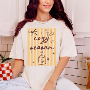 May include: Cream-colored t-shirt with a graphic that reads "cozy season" in brown script. The design includes yellow vertical stripes, a bow, mushrooms, a leaf, stars, and a mug with a leaf. The shirt is a relaxed fit.
