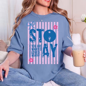 May include: A blue t-shirt with the word "STAY" in large blue letters, set against pink and white vertical stripes. The shirt also features the words "YOU ARE WANTED LOVED WORTHY" and small floral and bow graphics.