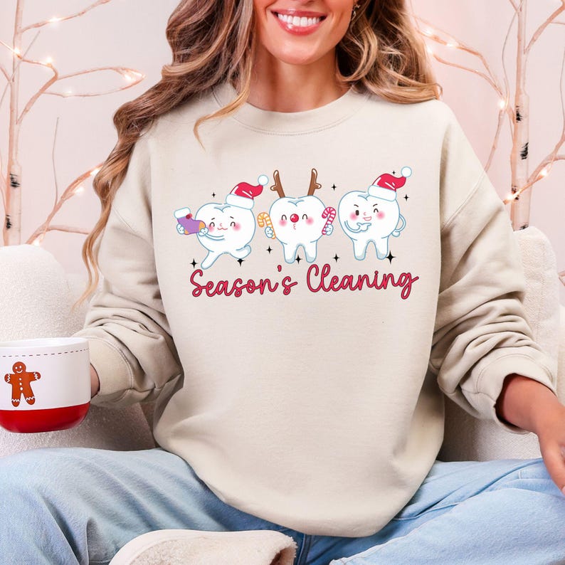 Puede incluir: Una sudadera beige con un gr&aacute;fico de tres dientes de dibujos animados con gorros de Pap&aacute; Noel y objetos festivos. El texto "Season's Cleaning" est&aacute; en escritura rosa debajo. Una taza blanca con un hombre de jengibre est&aacute; en primer plano.
