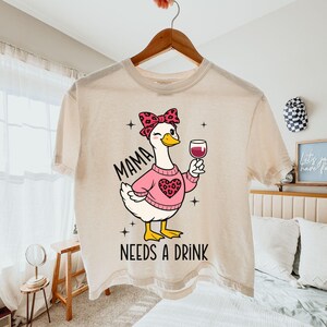 May include: Cream-colored t-shirt featuring a cartoon goose wearing a pink sweater with a heart design and a bow. The goose is holding a glass of red wine. Text on the shirt reads "MAMA NEEDS A DRINK".