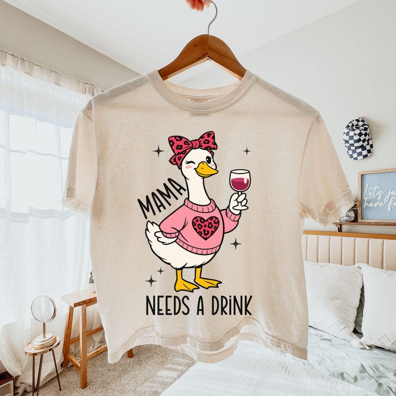 May include: Cream-colored t-shirt featuring a cartoon goose wearing a pink sweater with a heart design and a bow. The goose is holding a glass of red wine. Text on the shirt reads "MAMA NEEDS A DRINK".