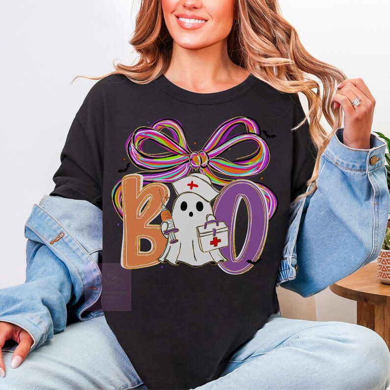 Pu&ograve; includere: Felpa nera con un fiocco colorato e la scritta "BOO" in lettere arancioni e viola. La "O" &egrave; un fantasma con un cappello da infermiera che tiene una siringa e una borsa medica. Design a tema Halloween.
