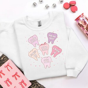 K&ouml;nnte beinhalten: Wei&szlig;es Sweatshirt mit einem Aufdruck von rosa und lila Cartoon-Z&auml;hnen, jeder mit einem zahnmedizinischen Spruch. S&auml;tze wie "Floss On" und "Sweet Tooth" sind zu sehen. Das Sweatshirt ist von der Marke Gildan. Dekorative Elemente sind verstreut.