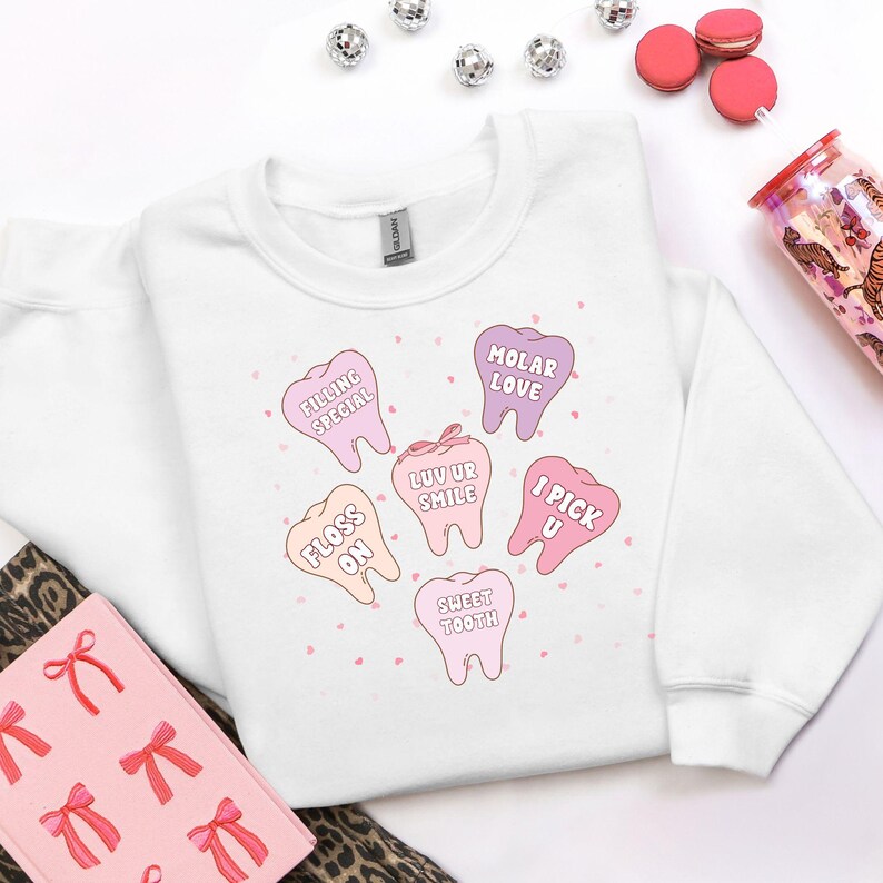 K&ouml;nnte beinhalten: Wei&szlig;es Sweatshirt mit einem Aufdruck von rosa und lila Cartoon-Z&auml;hnen, jeder mit einem zahnmedizinischen Spruch. S&auml;tze wie "Floss On" und "Sweet Tooth" sind zu sehen. Das Sweatshirt ist von der Marke Gildan. Dekorative Elemente sind verstreut.