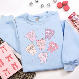 K&ouml;nnte beinhalten: Hellblaues Sweatshirt mit einem Aufdruck von Zeichentrickz&auml;hnen mit S&auml;tzen wie "Filling Special" und "Sweet Tooth". Das Bild enth&auml;lt rosa Makronen und einen Becher mit Muster.