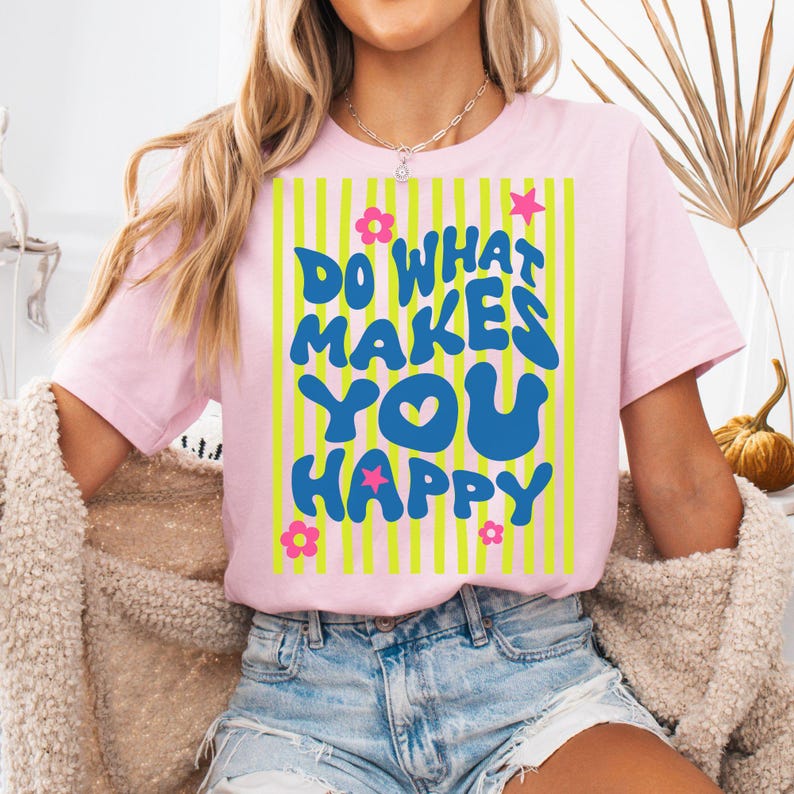 May include: Pink t-shirt with the text "DO WHAT MAKES YOU HAPPY" in blue, with a yellow and pink striped background. The design includes pink flowers and a pink star. The shirt is a short-sleeved crew neck.